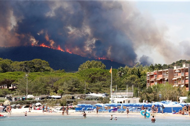 Pessoas se banham na praia em Bormes-les-Mimosas, no departamento de Var, na França, enquanto as chamas do incêndio florestal queimam no alto de uma colina
