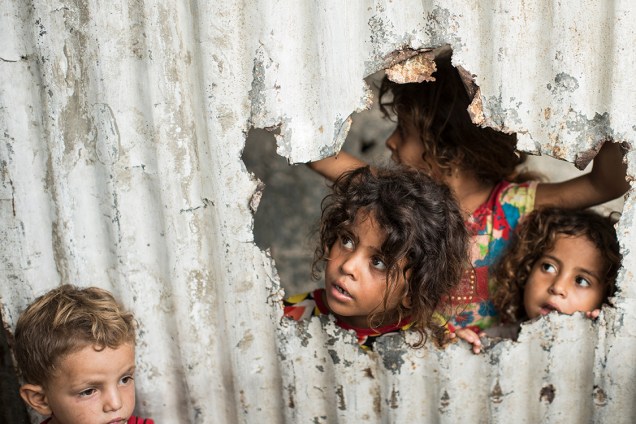 Crianças observam por um buraco em uma parede metálica ao lado de onde moram em Gaza  - 08/08/2017