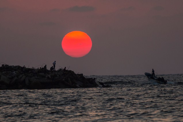 Um barco passa ao lado de uma costa rochosa onde palestinos observam o pôr do sol - 14/08/2017