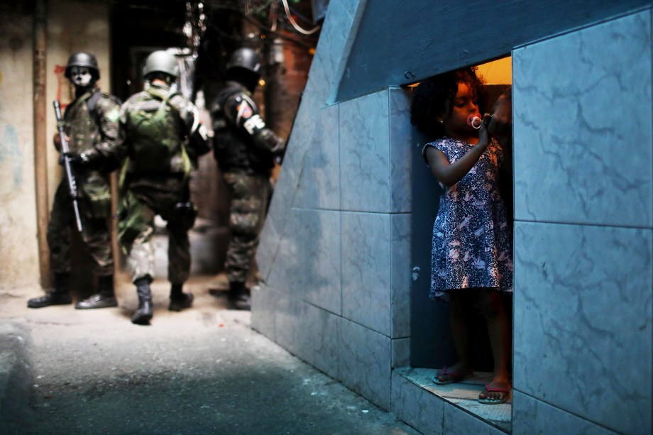 Uma garota é fotografada em uma porta sob uma escada enquanto ao seu redor policiais do Exército patrulham a Favela da Rocinha, durante operação no Rio de janeiro - 25/09/2017