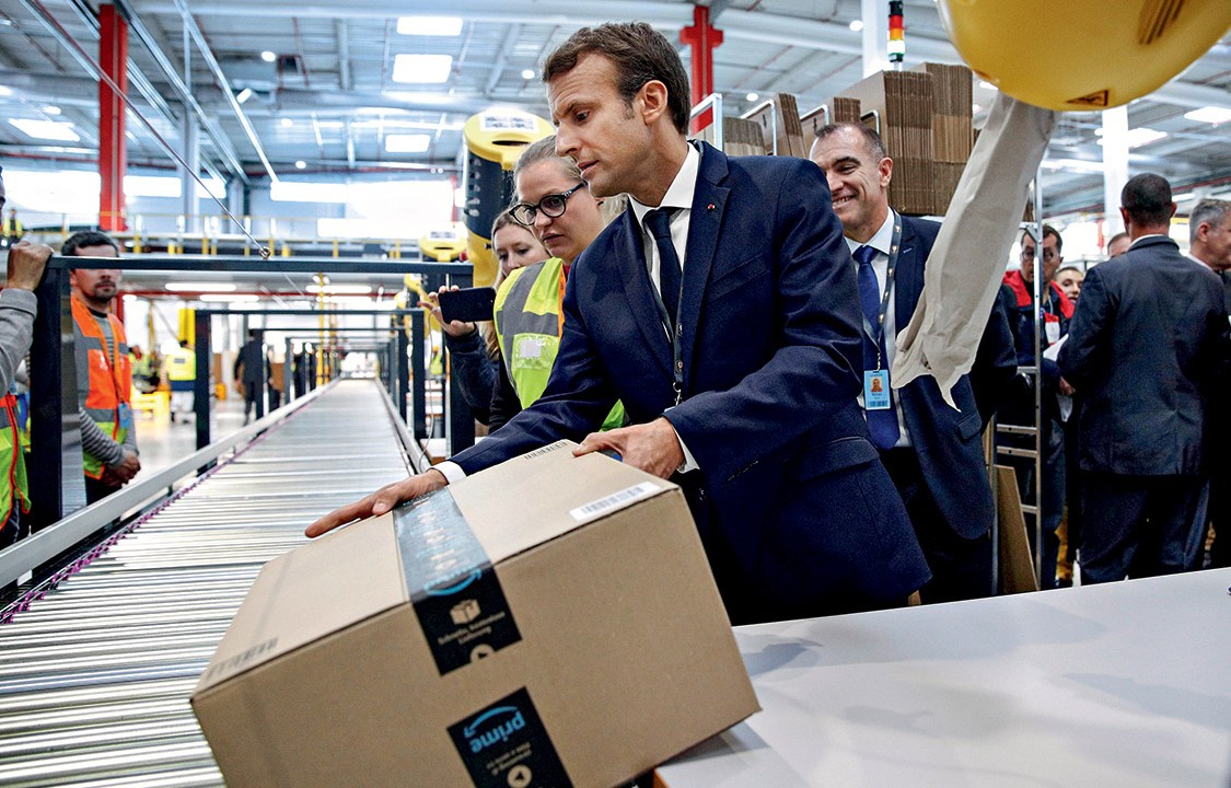 French President Emmanuel Macron (C), flanked by an employee (L) and Amazon French Operations Director Ronan Bole (R), moves a package during a visit at the Amazon factory in Boves, near Amiens, France, October 3, 2017. REUTERS/Yoan Valat/Pool