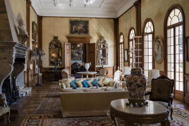 Janelas abobadadas, lareira, espelhos e as almofadas que descansam sobre o sofá, decoram o interior de uma das salas de estar da mansão Vila dos Cedros no estilo do século XIX