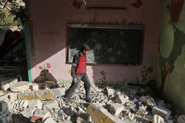 Menino sírio caminha sobre os escombros de sua escola na cidade sitiada de Saqba,  um dia após ataques aéreos do governo - 09/11/2017