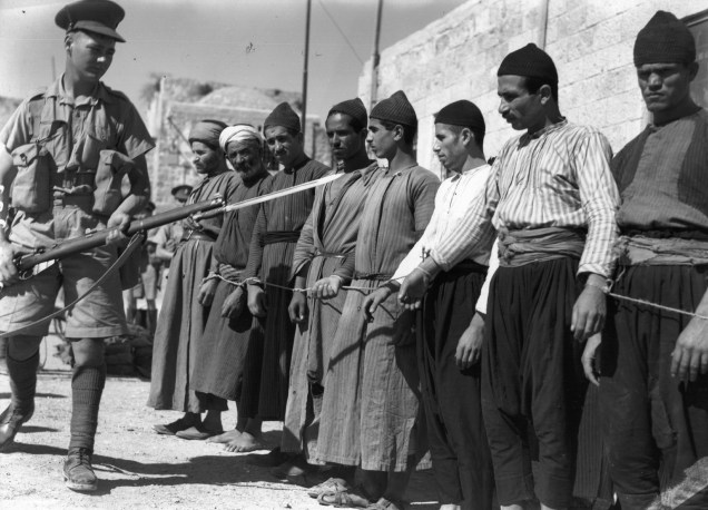 Um grupo de prisioneiros árabes na Cidade Velha de Jerusalém (durante o mandato britânico da Palestina) é escoltado por um soldado armado com um rifle e baioneta