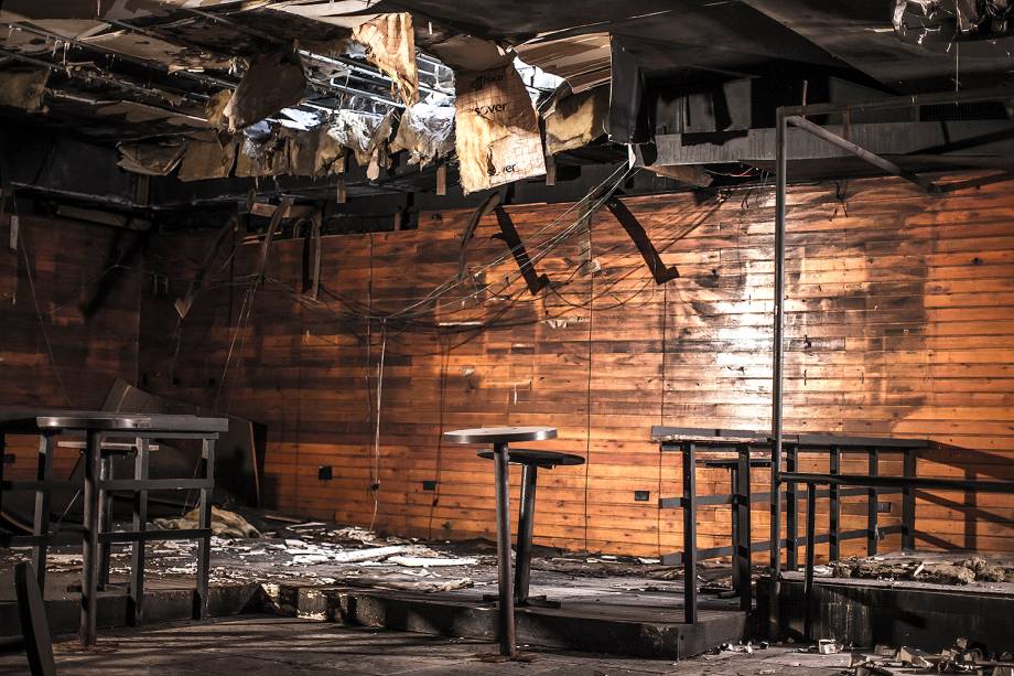 Parte do pub da boate. Mesinhas, antes fixas no chão, encontram-se tortas e, algumas, derrubadas após 5 anos da tragédia. As intempéries do clima abriram o buraco no teto, enfraquecido pelas chamas