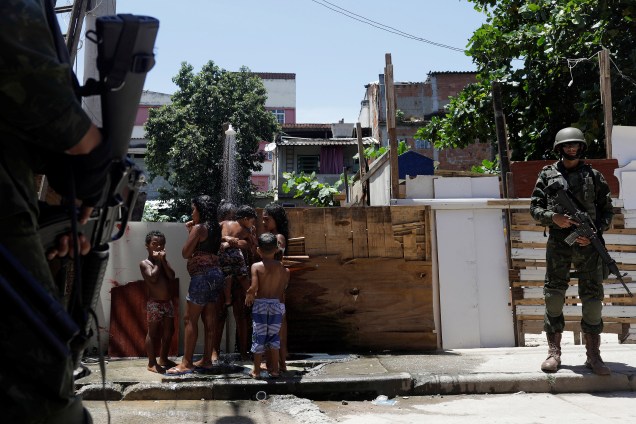Mulheres e crianças tomam uma ducha em um beco da favela de Manguinhos, no Rio de Janeiro, enquanto policiais do exército patrulham a região em operação contra o tráfico de drogas na região - 18/01/2018