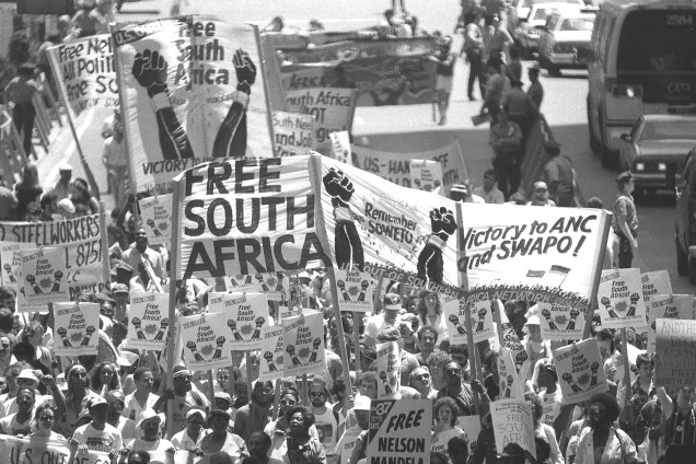 Manifestantes exibem faixas e cartazes durante protesto contra as políticas raciais implantadas durante o Apartheid, na África do Sul - 14/06/1986