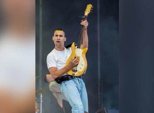Homem branco de camiseta branca e calça jeans clara tocando guitarra amarela em palco, com expressão concentrada e boca aberta, braço esquerdo levantado segurando o braço do instrumento