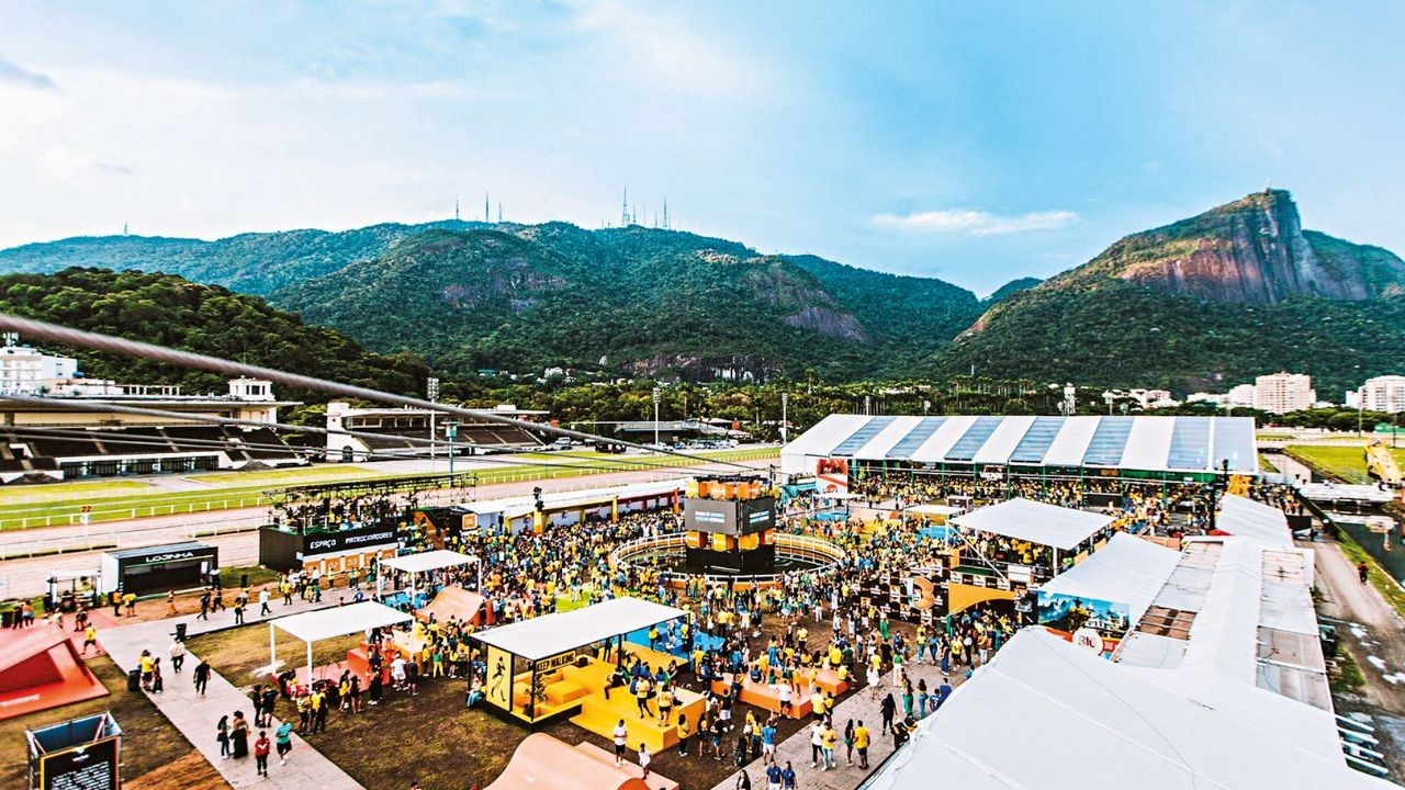 Vista aérea de um evento ao ar livre com multidão, tendas brancas e amarelas, e um palco central, em frente a uma pista de corrida e montanhas verdes sob céu azul