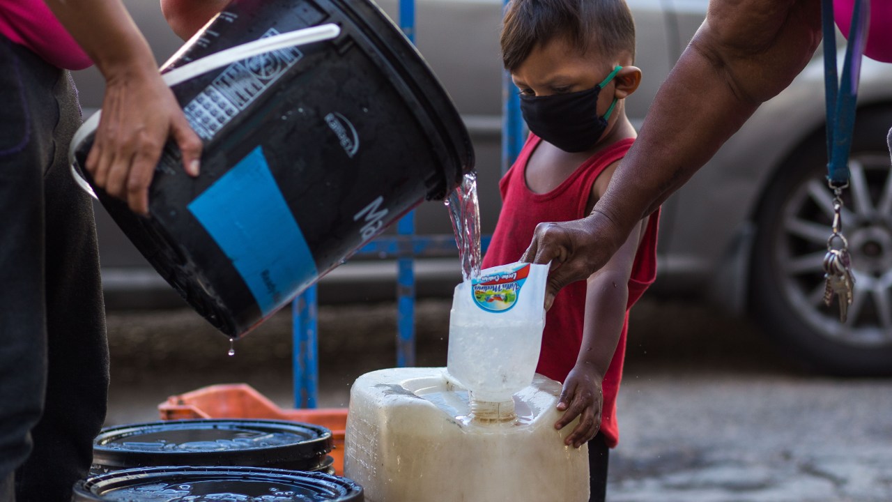 Criança com máscara observa coleta de água em recipientes precários em Caracas: crise humanitária e festas nababescas -27/03/2020