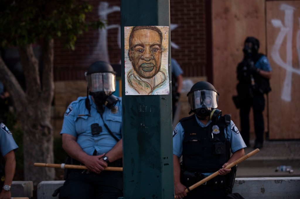 Um retrato de George Floyd pendurado em um poste de luz de rua enquanto policiais vigiam a 3ª Delegacia em Minneapolis, Minnesota