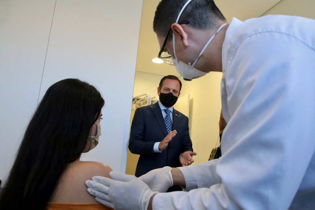 Uma voluntária recebe o primeiro teste da potencial vacina contra o coronavírus Sinovac, da China, de um profissional de saúde, enquanto é observada por João Doria, no Hospital das Clinicas, em São Paulo