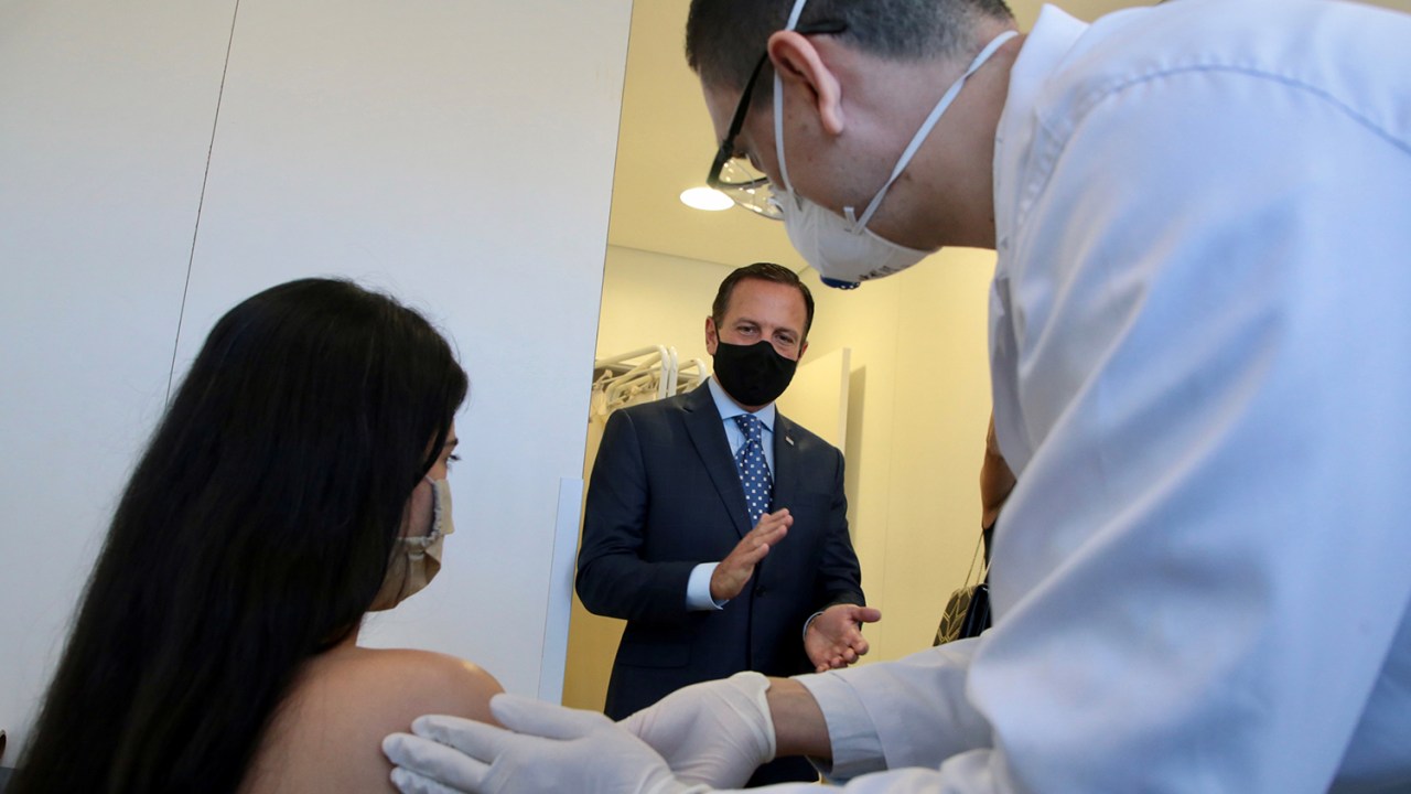Uma voluntária recebe o primeiro teste da potencial vacina contra o coronavírus Sinovac, da China, de um profissional de saúde, enquanto é observada por João Doria, no Hospital das Clinicas, em São Paulo