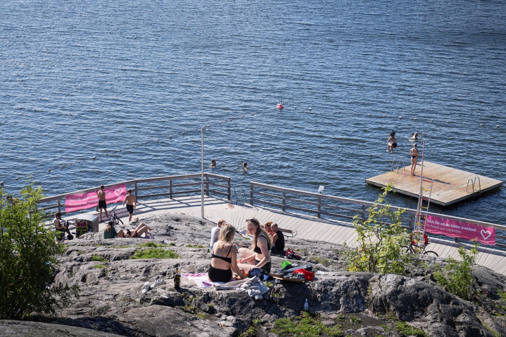 Pessoas socializam em um dia ensolarado de verão no lago Malaren, enquanto uma placa de incentivo ao distanciamento físico está pendurada em uma cerca em Estocolmo, Suécia - 31/07/2020