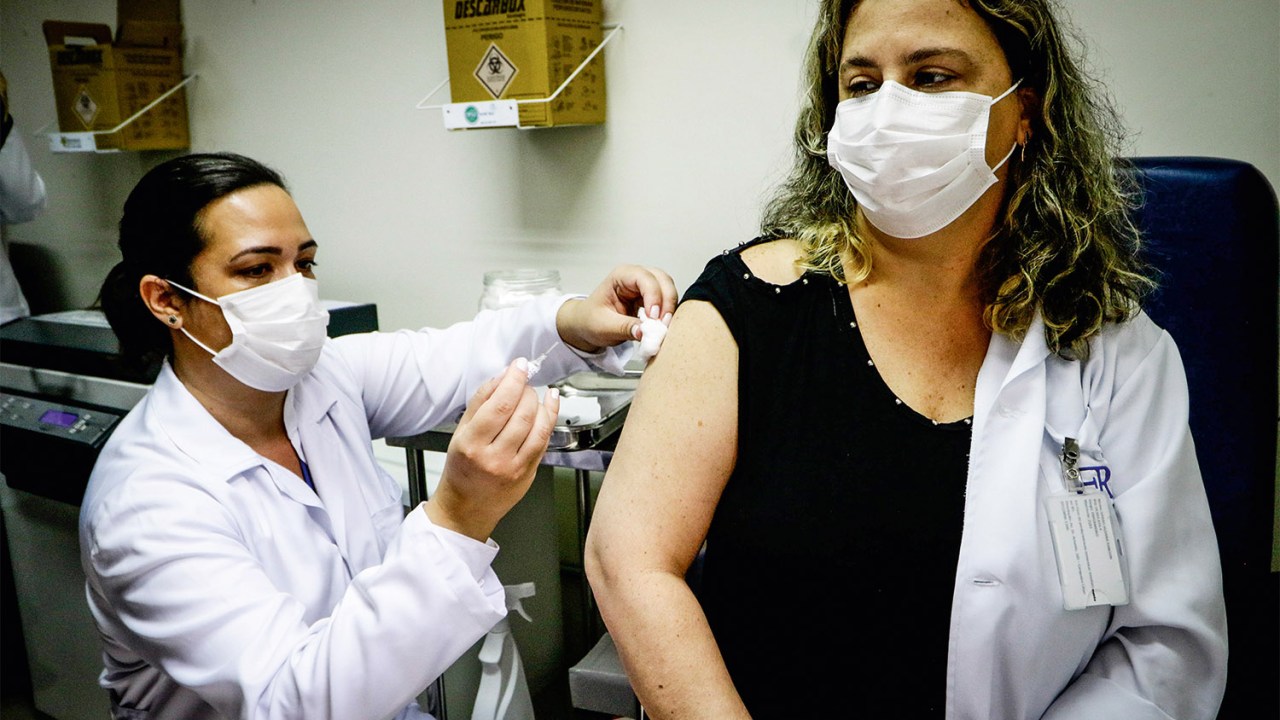 CoronaVac: imunizante da Sinovac. Na imagem uma médica da linha de frente ao combate do Coronavírus é vacinada. Uma enfermeira, que também usa máscara, aplica a vacina. Ambas usam máscaras de proteção.