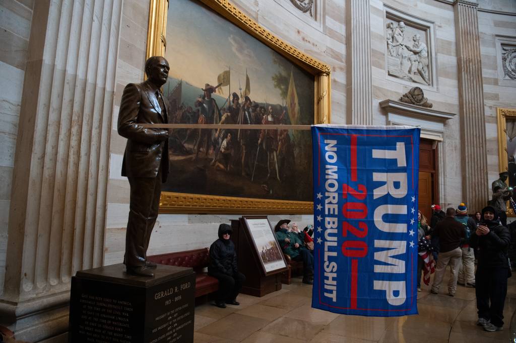 Estatátua adornada com bandeira de Donald Trump durante invasão ao Capitólio, quadro ao fundo em exposição na Rotunda