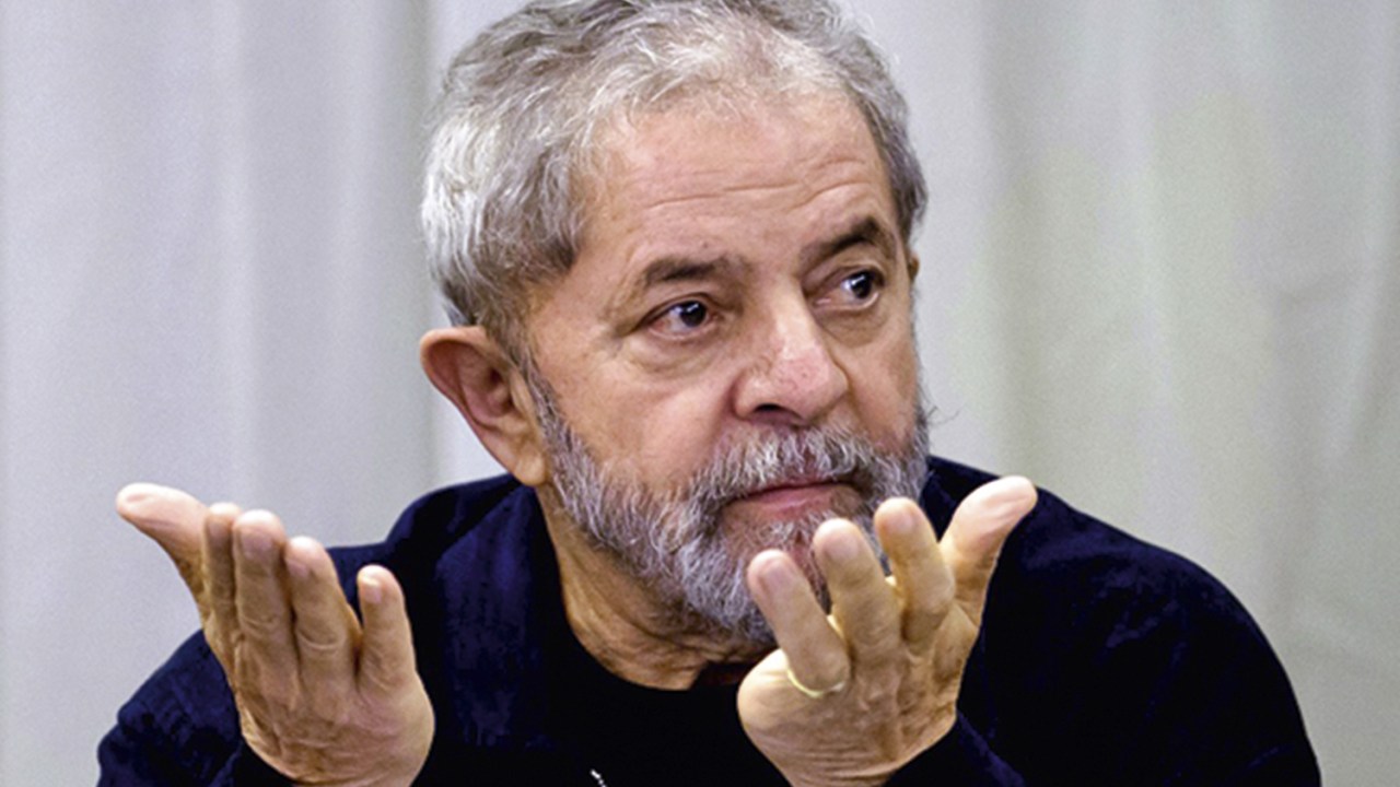 Defesa de Lula - Luiz Inácio Lula da Silva. Na imagem o ex-presidente Lula sentado com as mãos levantadas.
