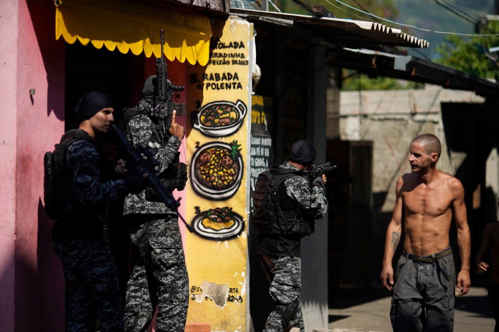 A local resident looks at Rio's Civil Police officers taking position during a police operation against drug traffickers at the Jacarezinho favela in Rio de Janeiro state, Brazil, on May 06, 2021. - A civil police officer and 22 suspects were killed during a police operation against drug traffickers in the Jacarezinho favela, Rio de janeiro on April 2021. The incident began with an exchange of fire at a metro station in the area which is in the North Zone of Rio de Janeiro. (Photo by MAURO PIMENTEL / AFP)