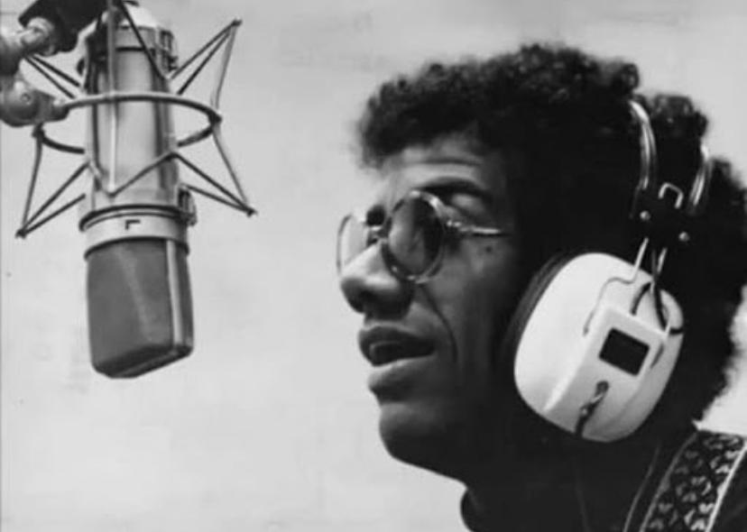 A imagem mostra Jorge Ben Jor na década de 70, ao microfone, com um fone no ouvido