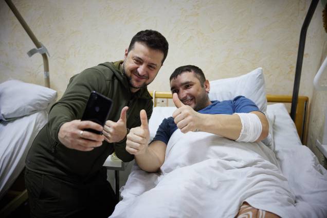 O presidente ucraniano Volodymyr Zelensky tira uma selfie com um homem ferido deitado em uma cama durante uma visita a um hospital militar após combates na região de Kiev -