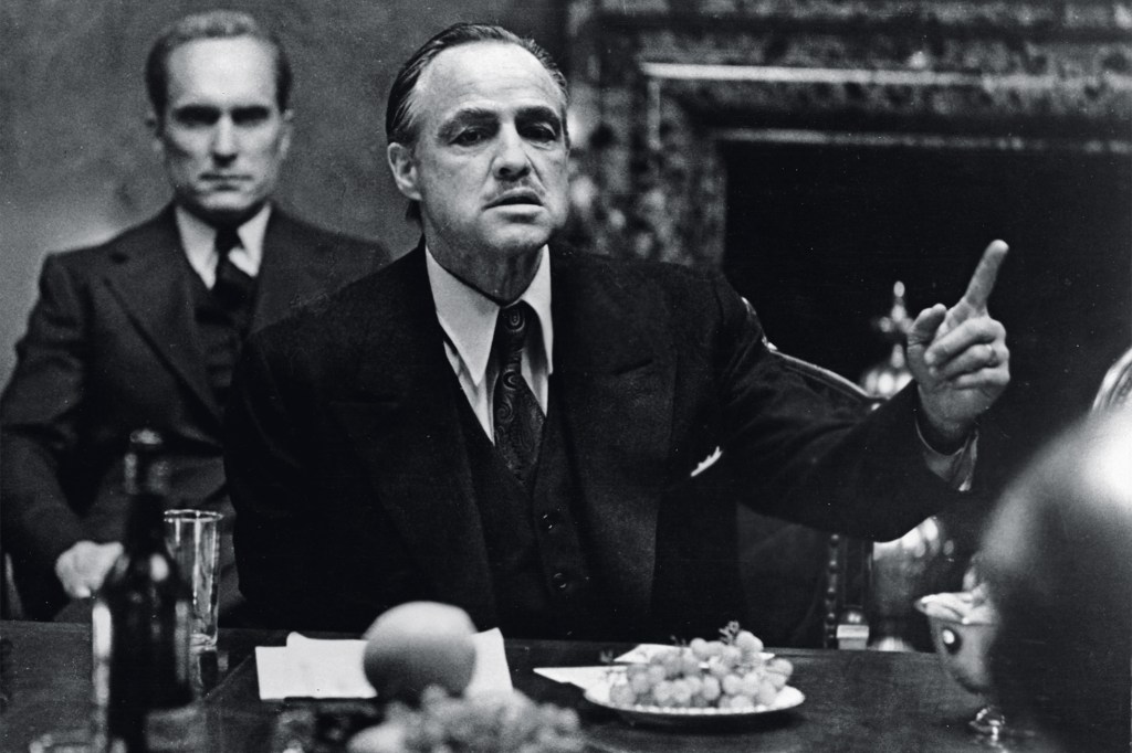 American actor Marlon Brando gestures at a table while American actor Robert Duvall sits behind him in a still from the film, 'The Godfather,' directed by Francis Ford Coppola and based on the novel by Mario Puzo. (Photo by Paramount Studios/Courtesy of Getty Images)