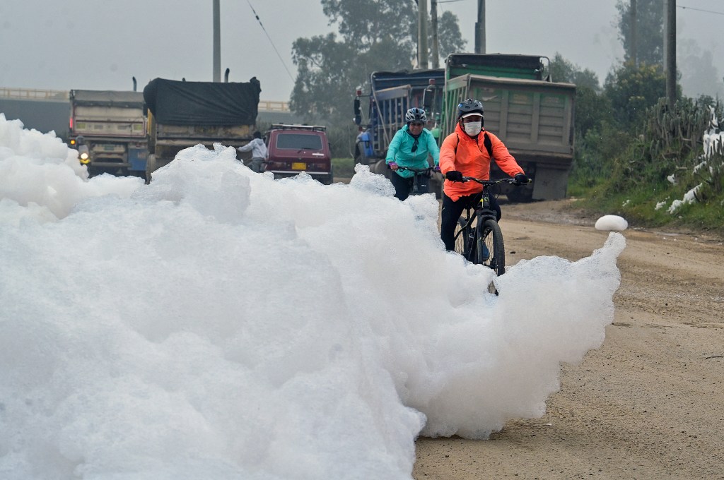 Cyclists ride by pungent foam which formed in a polluted river and invaded the Los Puentes neighbourhood, in Mosquera, west of Bogota, on April 26, 2022. (Photo by Juan BARRETO / AFP)