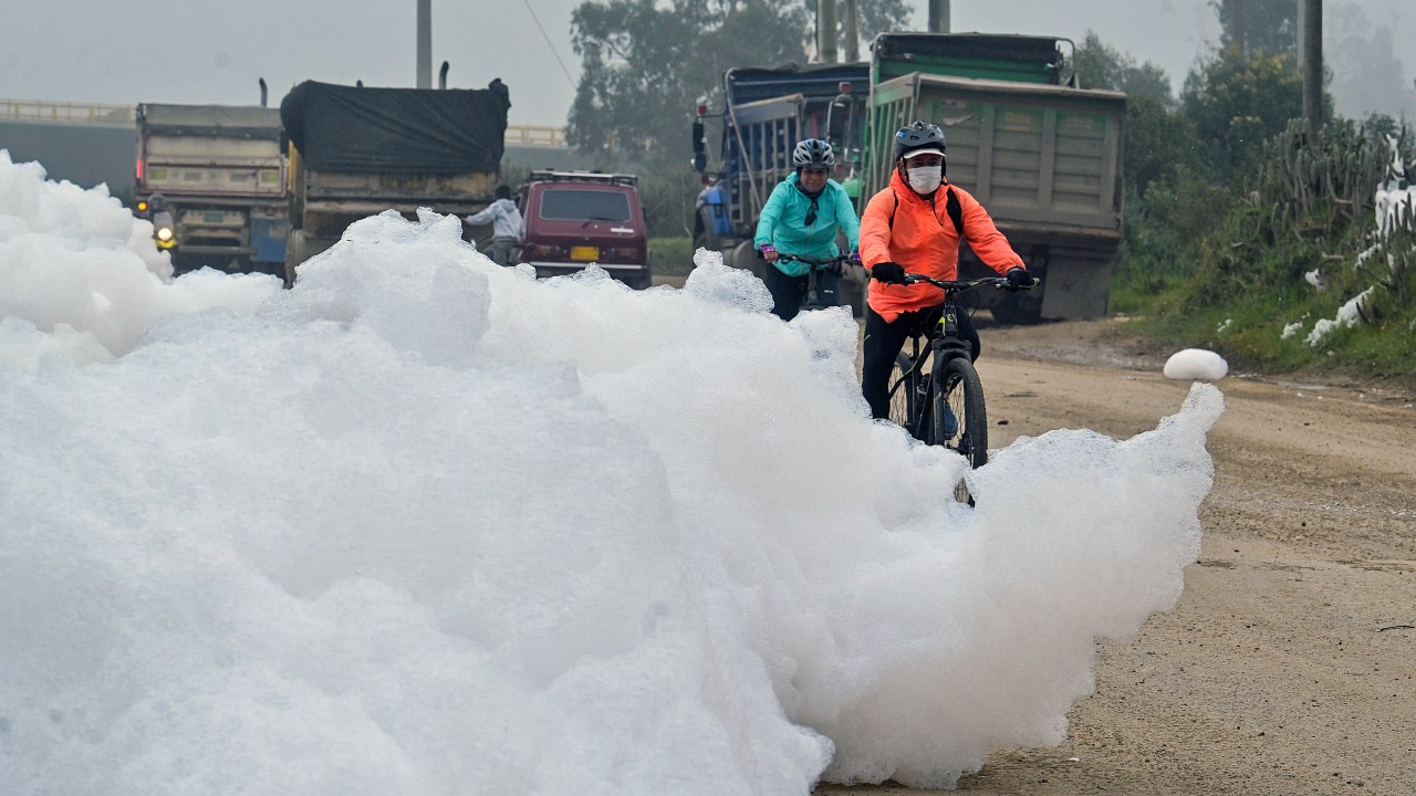 Cyclists ride by pungent foam which formed in a polluted river and invaded the Los Puentes neighbourhood, in Mosquera, west of Bogota, on April 26, 2022. (Photo by Juan BARRETO / AFP)