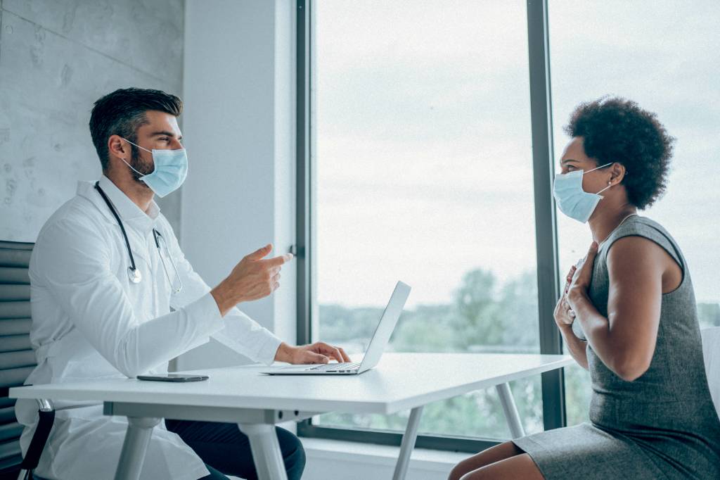 Young male doctor consulting patient inside his office at the hospital. African-american woman sitting opposite the doctor in his office and discussing her medical results. A patient and a male doctor meet in a medical clinic while wearing protective face masks to avoid the transfer of viruses.