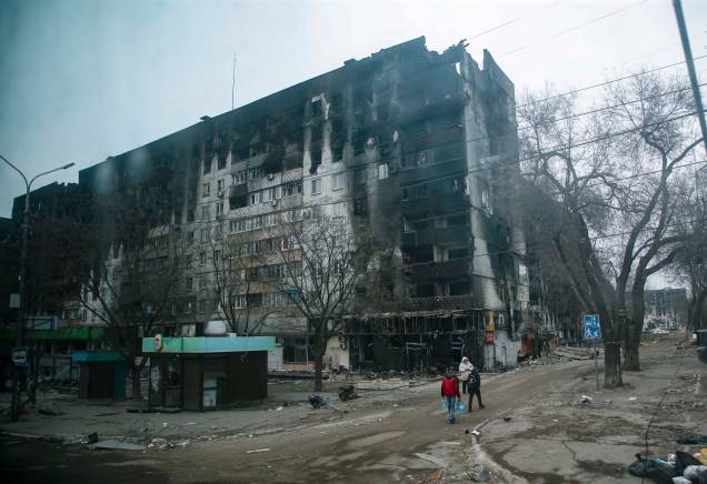 Pessoas carregando água próximo `a um prédio de apartamentos devastado e queimado no centro de Mariupol, Ucrânia, 12 de abril de 2022, pelo exército russo.