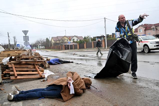 Um corpo de um homem civil com as mãos amarradas nas costas está na rua enquanto um trabalhador comunitário prepara um saco plástico para carregá-lo para um carro na cidade de Bucha, na Ucrânia -