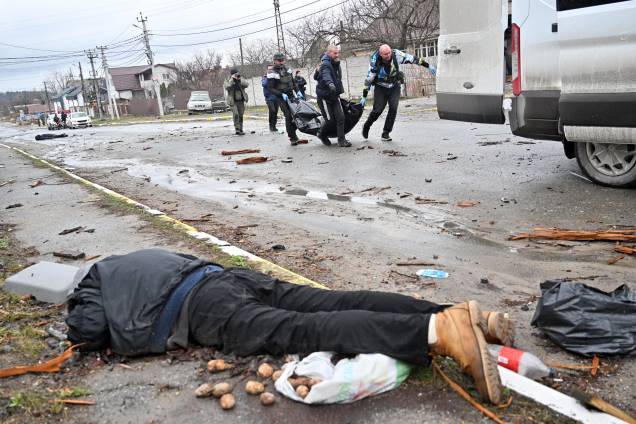 Trabalhadores comunitários carregam um civil em um saco de corpo enquanto outro cadáver é visto no chão na cidade de Bucha, na Ucrânia -