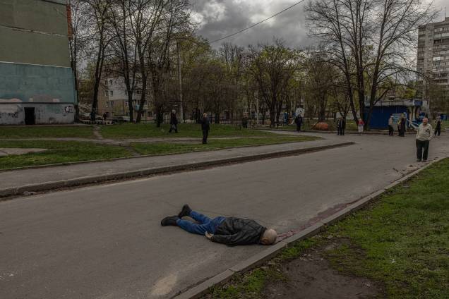 Pessoas olham para o corpo de um civil morto deitado em uma estrada na área residencial que foi atingida pelo bombardeio da artilharia russa, em Kharkiv, nordeste da Ucrânia, 19 de abril de 2022 -