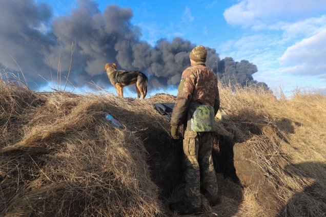 Um soldado ucraniano e seu cachorro em uma trincheira, olhando para a fumaça de um depósito de petróleo queimado após ser atingido por um míssil russo, próximo `a Kiev, Ucrânia, 27 de fevereiro de 2022.