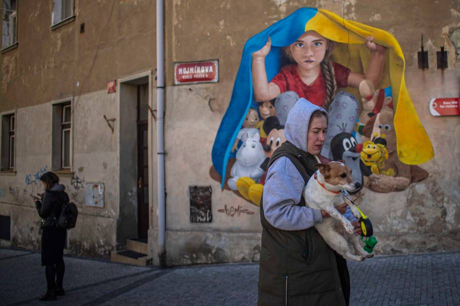 Refugiada ucraniana carregando um cachorro, passa por um mural que retrata uma criança se abrigando com figuras populares do entretenimento infantil sob a bandeira nacional da Ucrânia, criada pelo artista de rua 'ChemiS', em Praga , República Tcheca, 21 de março de 2022.
