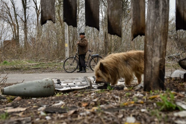 Um homem observa os destroços de um míssil enquanto um cachorro passa, Kukhari, Kiev Oblast, Ucrânia, 16 de abril de 2022.