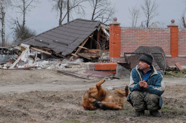 Um cachorro brinca ao lado de um morador local em frente a um prédio danificado na vila de Bohdanivka, próximo de Brovary, região de Kiev, Ucrânia, 12 de abril de 2022.