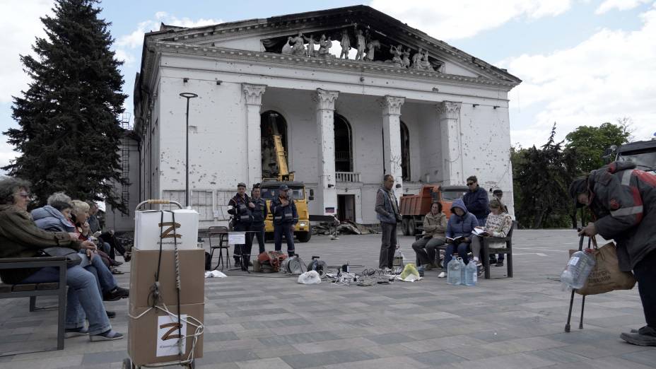 Moradores carregam seus celulares com um gerador, enquanto funcionários de emergências russas, limpa os destroços no teatro de drama Mariupol, parcialmente destruído na cidade de Mariupol em 10 de maio de 2022, em meio à ação militar russa em andamento na Ucrânia.