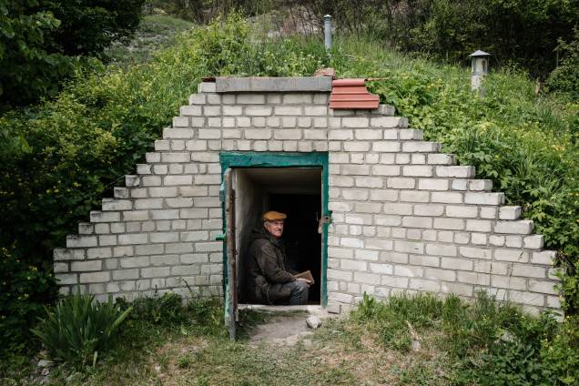 O professor de biologia ucraniano Oleksiy Polyakov, 84, lê o livro intitulado "200 Lugares Maravilhosos e Incríveis" na entrada de sua adega de vegetais transformada em abrigo em Sydorove, leste da Ucrânia, em 17 de maio de 2022.