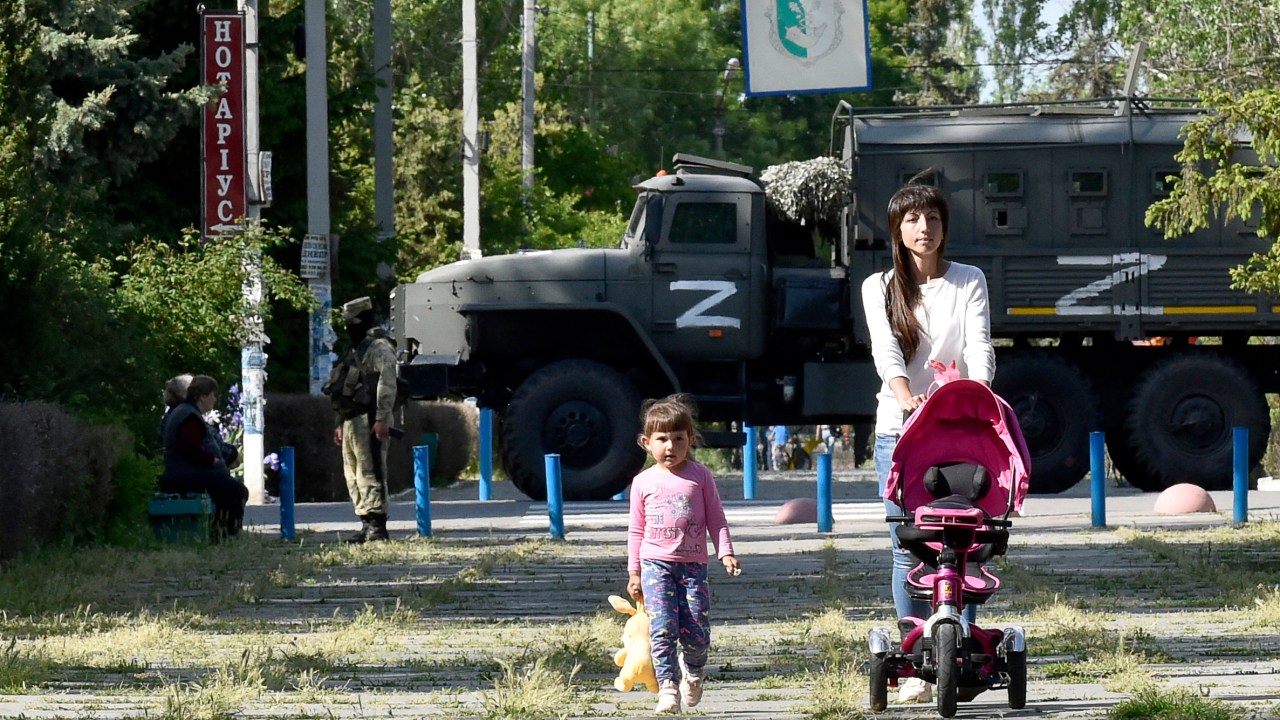 Esta foto tirada em 20 de maio de 2022 mostra uma mulher e uma criança andando em um parque enquanto militares russos patrulham a rua em Skadovsk, Kherson Oblast, em meio à ação militar russa em andamento na Ucrânia. (Foto de Olga MALTSEVA/AFP