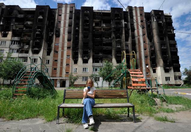 Uma menina olha para seu smartphone e aproveita o sol, ao fundo, prédios residenciais parcialmente destruídospelo bombardeio na cidade de Irpin, próximo da capital ucraniana de Kyiv, em 16 de junho de 2022.