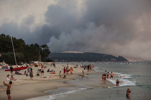 Pessoas na praia de Moulleau, ao fundo fumaça em virtude das altas temperaturas, ocasionando incêndios florestais em La Teste-de-Buch, vista de Arcachon, França, em 18/07/2022.