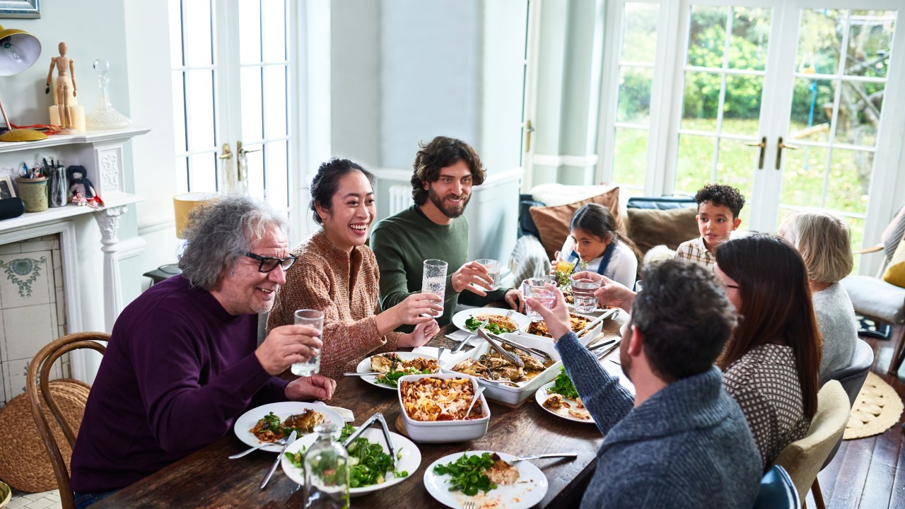 Group of mixed aged relatives toasting drinks at dining table enjoying home cooked meal talking and relaxing, celebration, healthy eating, meal time, domestic life