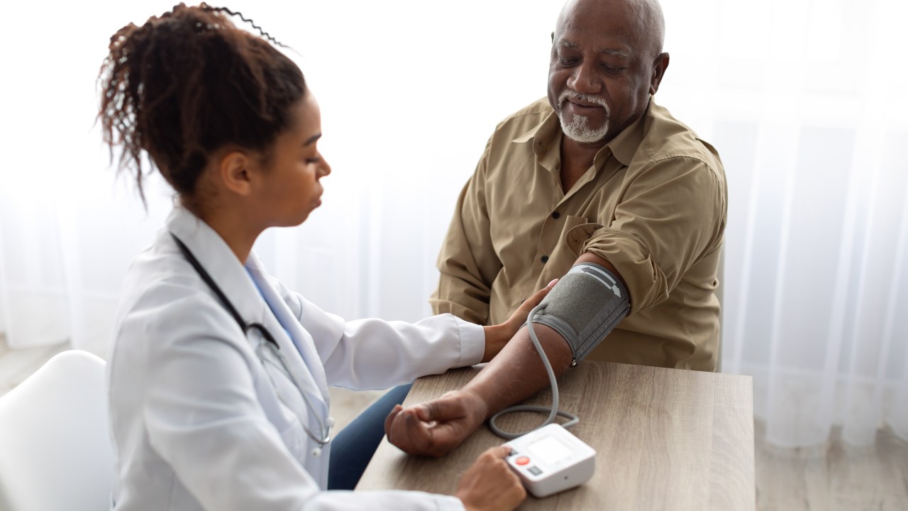 Hypertension In Older Age. Young Female Medical Worker Measuring Arterial Blood Pressure Of Senior Black Man Using Cuff, Patient Having Problems With Tension, Sitting At Table. Health Care Concept-letra de medico