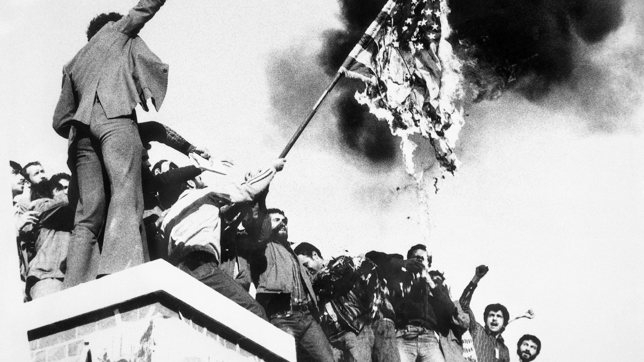 (Original Caption) 11/9/1979 Teheran, Iran: Demonstrators perched atop of the United States Embassy wall, burn an American flag, the fourth American flag to be burned since the students seized the embassy and more than 60 hostages Nov. 4th. demonstrators also burned an efigy of President Carter, Nov. 9th.