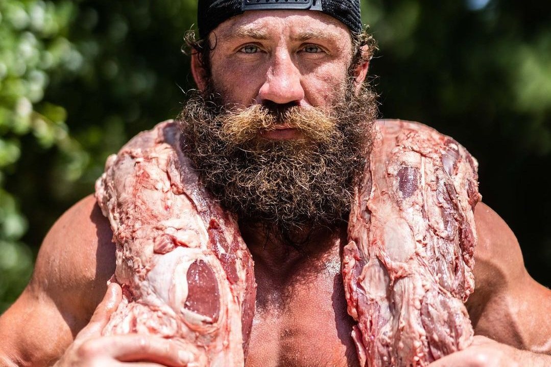 Homem musculoso e barbudo segurando uma peça de carne sobre suas costas, enquanto usa um boné virado para trás