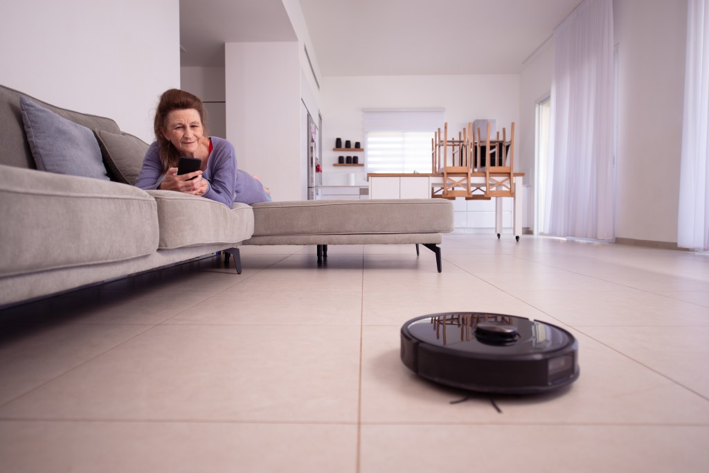 Relaxed senior housewife lying on a sofa, using a smartphone to control a robot vacuum cleaner. Robotic vacuum cleaner working, cleaning living room's tiled floor. Smart home and managing housework with a mobile device.