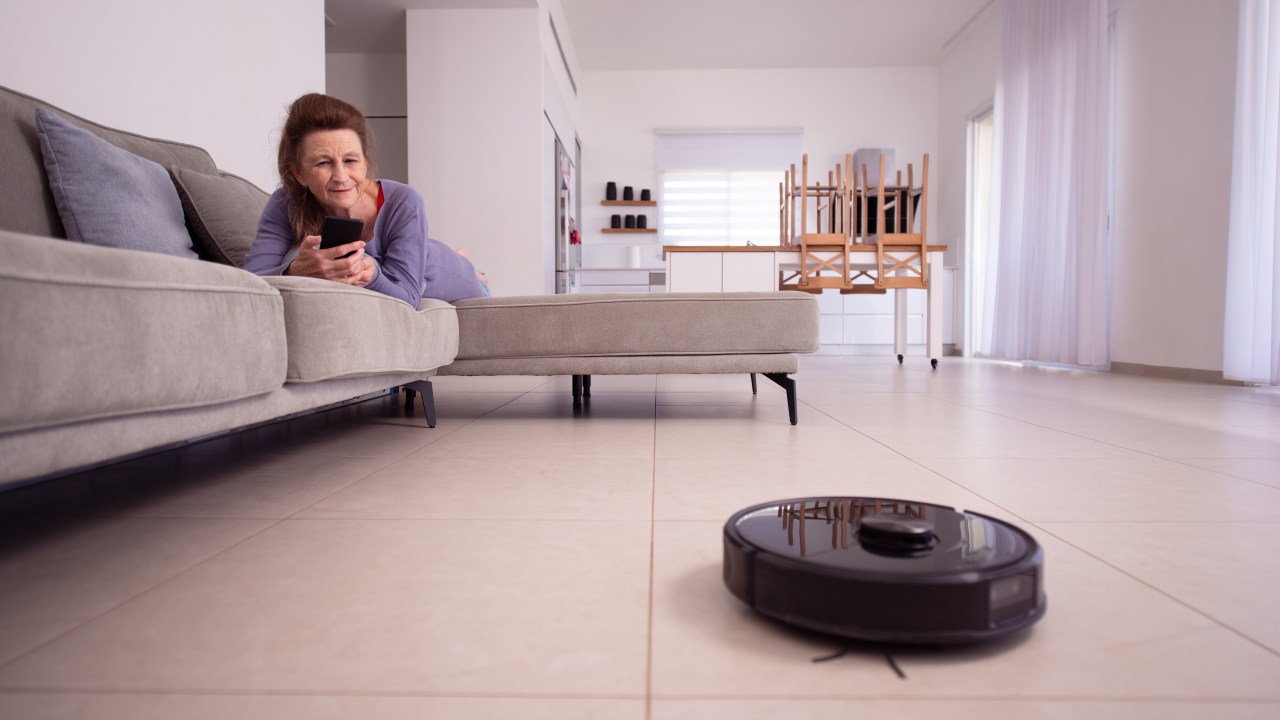 Relaxed senior housewife lying on a sofa, using a smartphone to control a robot vacuum cleaner. Robotic vacuum cleaner working, cleaning living room's tiled floor. Smart home and managing housework with a mobile device.