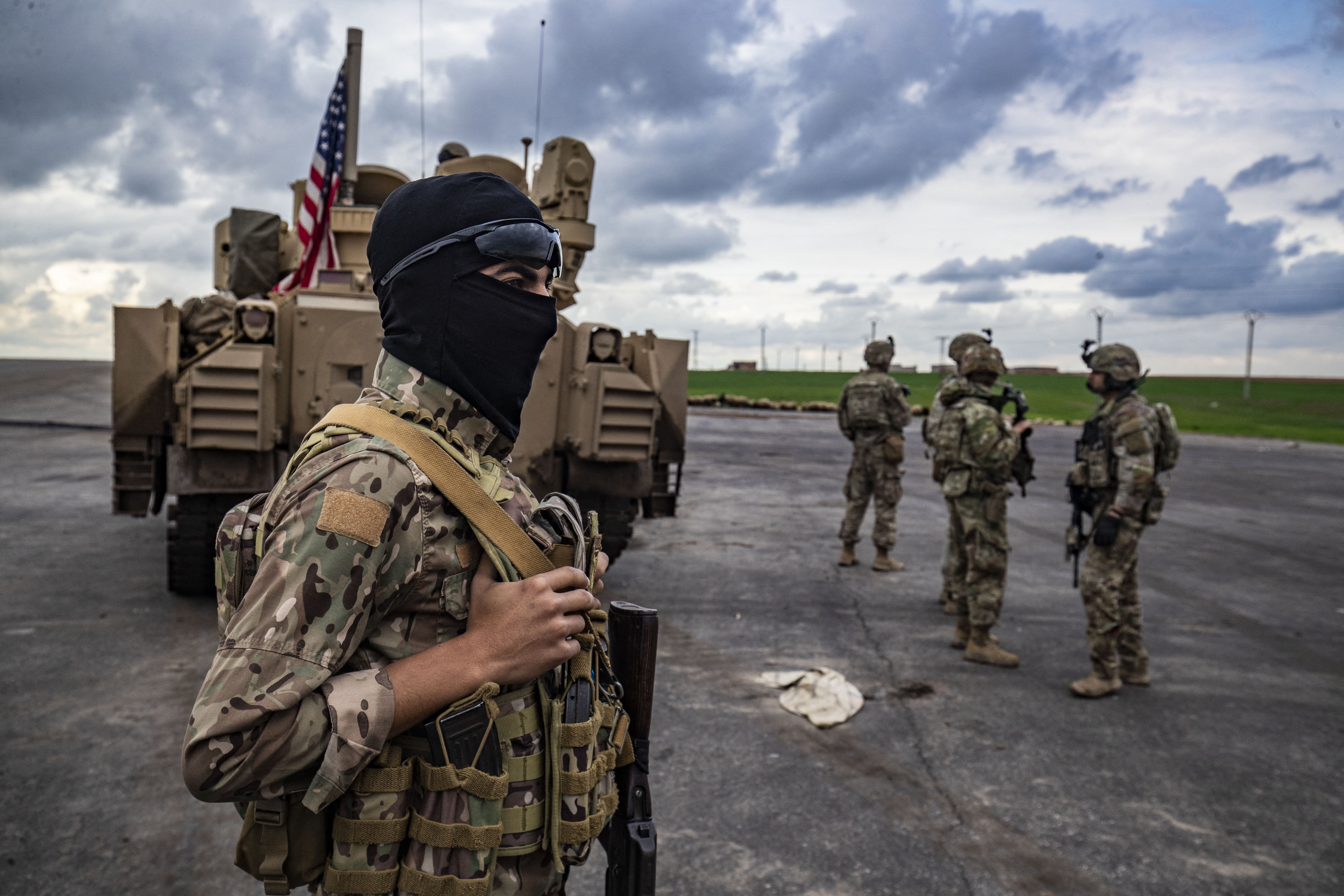 A member of the Syrian Democratic Forces (SDF) (L) and US soldiers are pictured near an armoured military vehicles are pictured on the outskirts of Rumaylan in Syria's northeastern Hasakeh province, bordering Turkey, on March 27, 2023. (Photo by Delil souleiman / AFP)