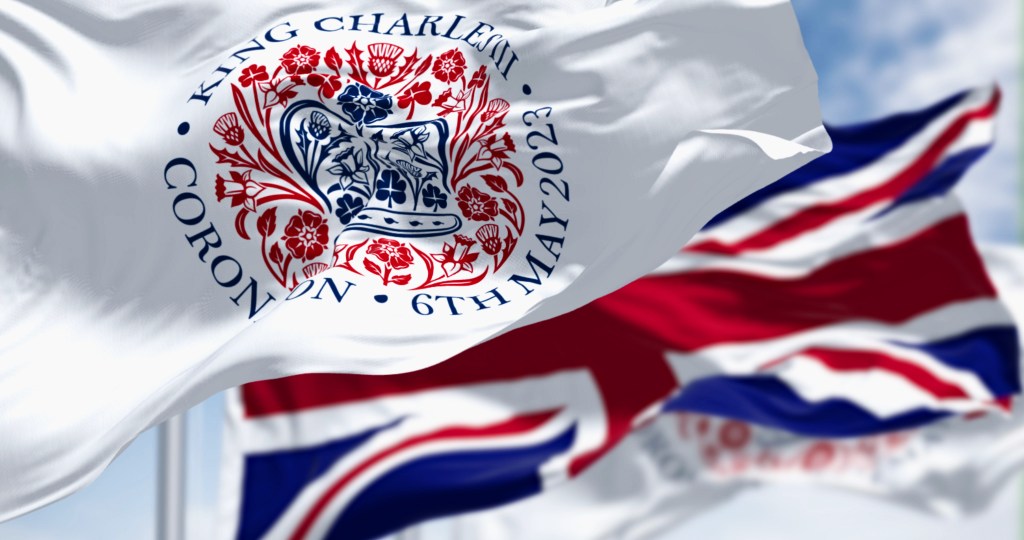 Flags with the emblem of the coronation of King Charles III and of UK waving. The emblem depicts flora of the four nations in the shape of St Edward’s Crown. 3d illustration render. Fluttering textile
