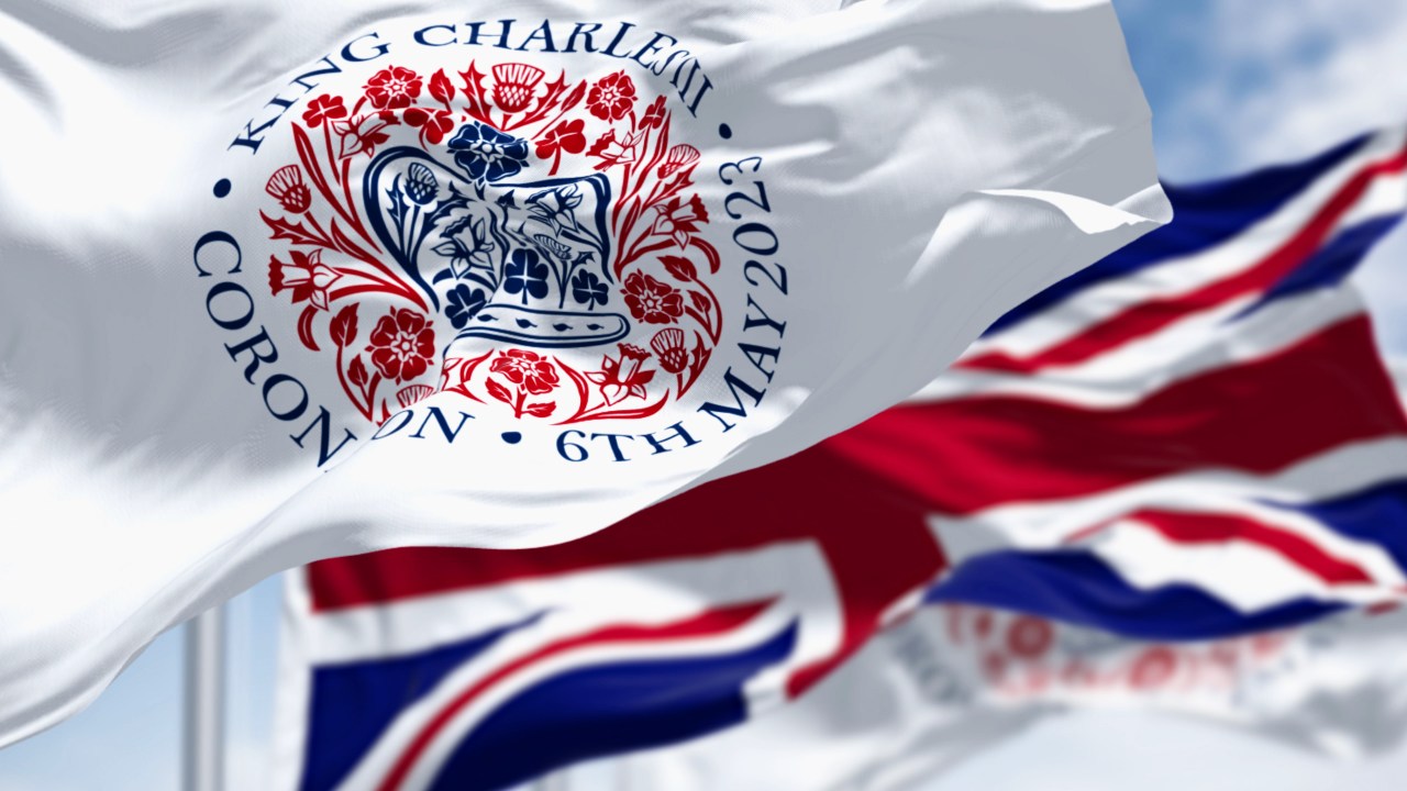 Flags with the emblem of the coronation of King Charles III and of UK waving. The emblem depicts flora of the four nations in the shape of St Edward’s Crown. 3d illustration render. Fluttering textile